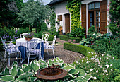 Round terrace with white seating area, bed with Hosta (Funkien), Geranium (Storchschnabel), hedge of Buxus (Buchs), bird bath, Pyrus salicifolia Pendula (Willow-leaved pear), Humulus lupulus 'Aureus' (Golden hop) - by the house
