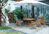 Sitzgruppe unter Sonnenschirm auf Terrasse am Haus, Brennholz-Stapel, Datura syn. Brugmansia (Engelstrompete) in der Ecke