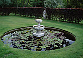 Round pond basin with Nymphaea (water lilies) in the lawn, stone figure stands in hedge of Fagus purpurea (copper beech)