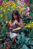 Mädchen mit frisch geerntetem Gemüse im Korb, dahinter hohe Blühpflanzen