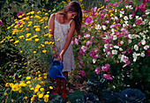 Gießen im Garten: Mädchen giesst Kohl (Brassica), Anthemis (Färberkamille) und Cosmos (Schmuckkörbchen) im Hintergrund