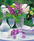 Purple and white syringa in tall glass vase, flowers, carafe, wine glass