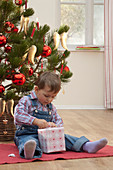 Kleiner Junge mit Geschenk sitzt vor Weihnachtsbaum