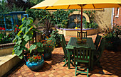Grüner Tisch und Stühle auf Terrakotta-Terrasse mit Wasserspiel dahinter und Calceolaria F1 Hybrid 'Sunset' und Paulownia in Töpfen