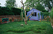 Spielbereich für Kinder mit blauem Gartenhaus, Tafel, Hängematte, Rasenkrokodil und altem Baum
