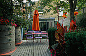VIEW TOWARDS FLAT with Blue Café CHAIRS, Pink WOODEN TABLE, Orange Parasol, DECKING, Orange Canna AND GALVANISED TRAY PLANTED with SUCCULENTS: Designer: STEPHEN WOODHAMS