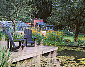 VIEW OVER LILY Pool TO WOODEN DECK with Adirondack CHAIRS. IN THE BACKGROUND ARE TWO GREENHOUSES. GREYSTONE Cottage, OXFORDSHIRE. Designer: DUNCAN HEATHER