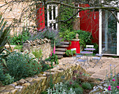 Blick auf die Rückseite des Hauses mit roter Wand, Stachys, Phormium, Metalltisch und Stühlen