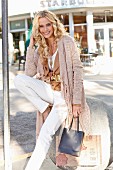 A blonde woman on a shopping street wearing a brown suede jacket, a cardigan with fringing and white trousers, holding a shopping bag