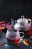 Autumn red hibiscus tea in glass cup and teapot standing on dark background with fall maple leaves and chestnuts