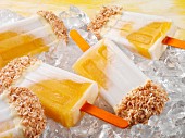 Mango and coconut ice lollies topped with white chocolate and desiccated coconut on a bed of ice cubes (close-up)