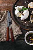 Sliced burrata served on round chopping board with pine nuts, sliced bread, basil and dried tomatoes in olive oil