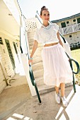 A brunette woman wearing a cropped top, a statement necklace, a pink tulle skirt, lace-up shoes and holding a clutch