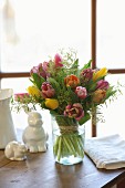 Luxuriant bouquet of tulips and delicate touch-me-not in glass vase next to china rabbits