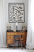 Candelabras and table lamp on old wooden desk below artwork on wall