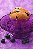A blueberry muffin on a small purple glass cake stand sitting on a purple crushed paper background