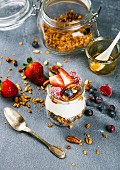 Yogurt oat granola with fresh berries, nuts, honey and mint leaves in glass jar on grey backdrop