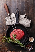 Raw meat Ribeye steak, seasonings and meat fork on cast iron frying pan on wooden background