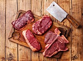 Different types of Raw fresh meat Steaks and meat cleaver on cutting board on wooden background