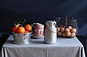 An arrangement of clementines, vintage coffee cups, an aluminium milk jug and eggs