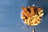 Chicken fries strips and legs with French fries in metal basket over blue wooden table with sea salt