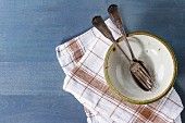 Empty ceramic bowl with vintage cutlery on kitchen checkered towel over blue wooden surface