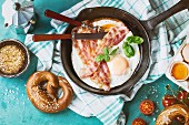 Breakfast with fried eggs and bacon in cast-iron pan, broken egg, tomatoes, mustard, pretzels and basil
