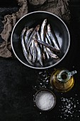 Lot of raw fresh anchovies fishes in black ceramic bowl with sea salt and bottle of olive oil for marinade