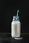Geöffnete Glasflasche mit Milch und Strohhalm vor schwarzem Hintergrund