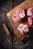 Raw osso buco meat on wooden cutting board with vintage steelyard, salt, pepper and rosemary over old wooden table