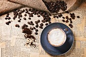 Blue ceramic cup of cappuccino with roasted coffee beans over old newspaper