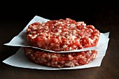 Two raw hand made beef burger patties stacked on white wax paper Dark wood background