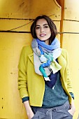 A brunette woman wearing a grey pullover, a yellow jacket and a scarf in front of a yellow background