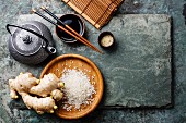 Raw white rice, green tea teapot, soy sauce and sushi chopsticks on gray stone slate background