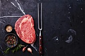 Rohes Black Angus Ribeye Steak mit Fleischgabel und Gewürzen auf dunklem Untergrund
