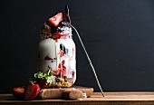 Yogurt oat granola with strawberries, mulberries, honey and mint leaves in tall glass jar on black backdrop