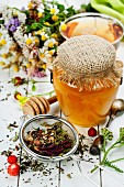Teestilleben mit Kräutertee im Sieb, Honigglas, Beeren und Blüten