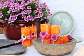 Orangefarbene Kerzen mit Blüten und Geschenkbändern dekoriert in rustikaler Schüssel