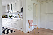 Pink chair next to open doorway leading into white country-house kitchen