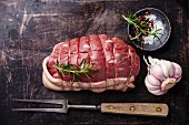 Raw roast beef, seasonings and meat fork on dark metal background