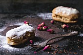 Eclairs with sugar powder, served with dry tea rose buds and vintage cutlery over dark background