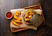 Black burger with sesame seed bun, meat, bacon, onion rings fries and potato wedges on dark wooden background