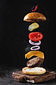 Flying ingredients for homemade burger on little wooden cutting board over dark background