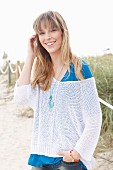 Blonde Frau in blauem T-Shirt, weißem Strickpullover und Jeans am Strand