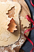 Cookie dough rolled out on a floured wooden board with a copper tree shape cookie cutter cutting out tree shape bluwe napkin and noel red ribbon