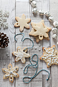 Overhead graphic shot of snowflake shaped christmas biscuits some docorated with white icing a sprinkles being threaded with blue and white bakers twine on a rustic whitewash wooden surface surrounded with Christmas decorations