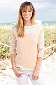 A blonde woman wearing a cream jumper and white trousers on the beach