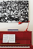 Sheet music and red lamp on red piano below large black and white photograph