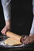 Female hands rolling out dough for pasta by wooden rolling pin over wooden kitchen table, powdering by flour