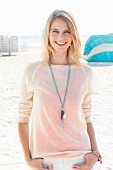 A blonde woman wearing a knitted jumper, white trousers and a long necklace at the beach